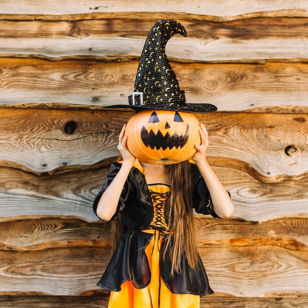 Girl in witch costume closing face by pumpkin with hat