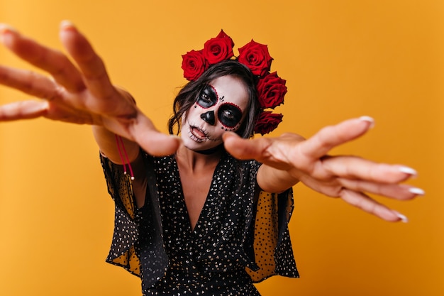 Girl with beautiful but frightening face art pulls hands towards camera like zombie. Portrait of unusual woman with red roses in her hair.