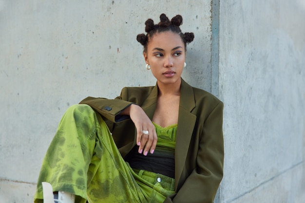 Free Photo girl with buns hairstyle vivid makeup dressed in green jacket and jeans poses against grey wall looks away being deep in thoughts rests after walking outside inn city