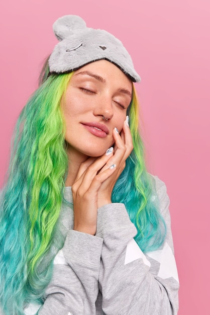 Free photo girl with closed eyes touches face gently sees sweet dreams while napping wears sleepmask on forehead comfortable pajama has dyed colorful hair isolated on pink wall