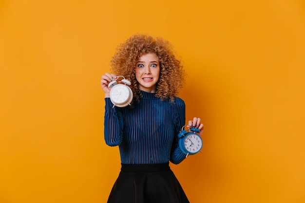 Free photo girl with curls dressed in blue sweater feels awkward holding two alarm clocks on yellow background