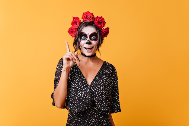 Girl with face painted for Halloween has new funny idea. Portrait of stylish young woman with roses in her hair.