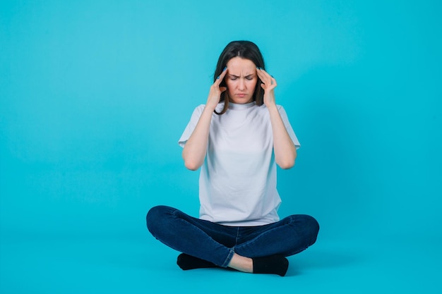 Free Photo girl with headache is holding ahnds on temples and sitting on floor on blue background