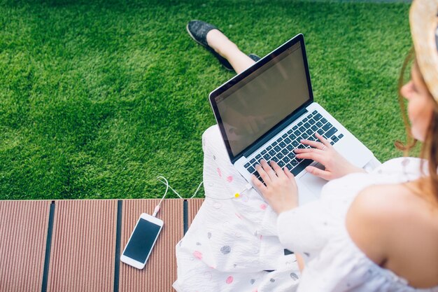 Girl with long hair in hat is sitting on planking floor. She wears white dress with naked shoulders. She is typing on a laptop on knees. View from above, focus on laptop