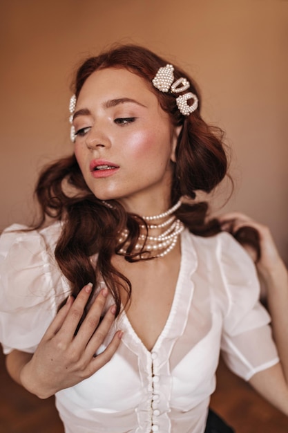 Free Photo girl with natural makeup posing on beige background lady in white blouse with pearl jewelry gently touches her body