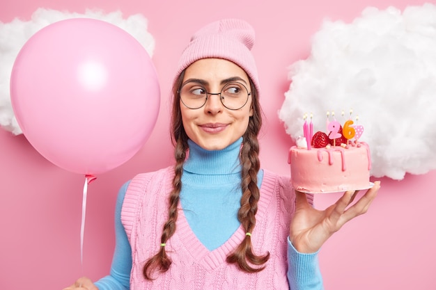 Free photo girl with pigtails looks dreamy concentrated aside wears round spectacles and casual clothes holds inflated balloon cake isolated on pink