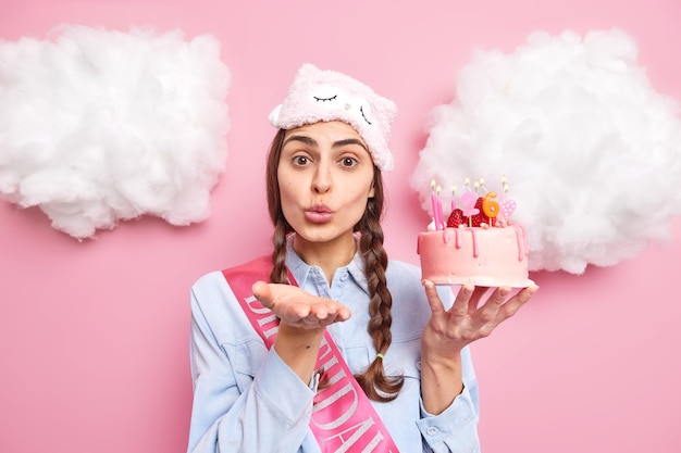 Free Photo girl with pigtails sends airkiss keeps lips folded keeps palm forward lips wears sleepmask shirt and ribbon holds tasty festive cake with burning candles isolated on pink 