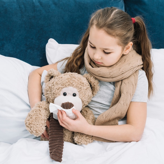 Girl with scarf around her neck covering the mouth of teddy bear with tissue paper