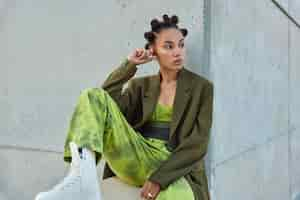 Free photo girl with trendy hairstyle dressed in green clothes looks away poses against urban grey wall considers something