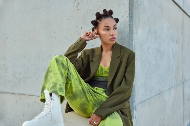 Free Photo girl with trendy hairstyle dressed in green clothes looks away poses against urban grey wall considers something