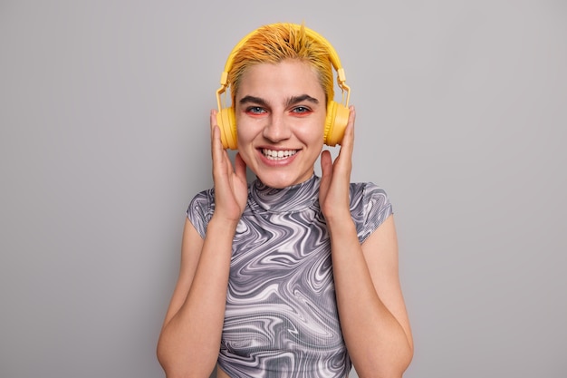 Free Photo girl with trendy yellow hairstyle bright makeup listens music in wireless headphones has cheerful mood enjoys favorite rock song dressed casually on grey