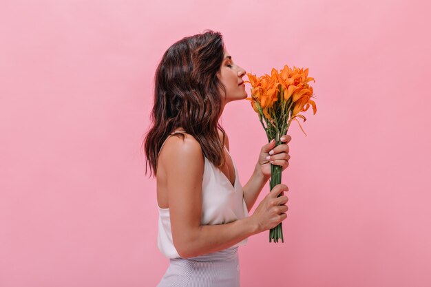 Girl with wavy hair enjoys scent of orange beautiful flowers. Dark-haired woman in light fashionable outfit sniffs large bouquet.