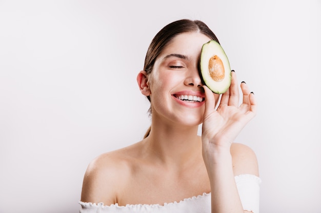 Girl without makeup holds half avocado, covering part of face. Pretty female model with dark hair leads healthy lifestyle.