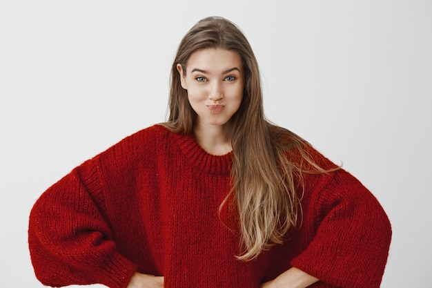 Girls making bet how long they can hold breath. Indoor shot of childish attractive feminine woman in stylish red loose sweater, holding hands on hips and sulking playfully 