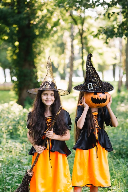 Free Photo girls in sorceress costumes holding broomstick and closing face by pumpkin