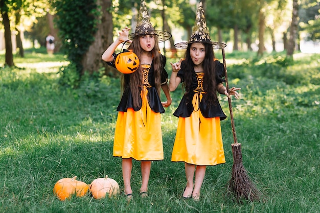 Free photo girls in witch costumes making faces, holding broomstick and pumpkin