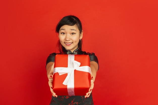 Giving giftbox. Happy Chinese New Year 2020. Asian young girl's portrait isolated on red background. Female model in traditional clothes looks happy. Celebration, holiday, emotions. Copyspace.