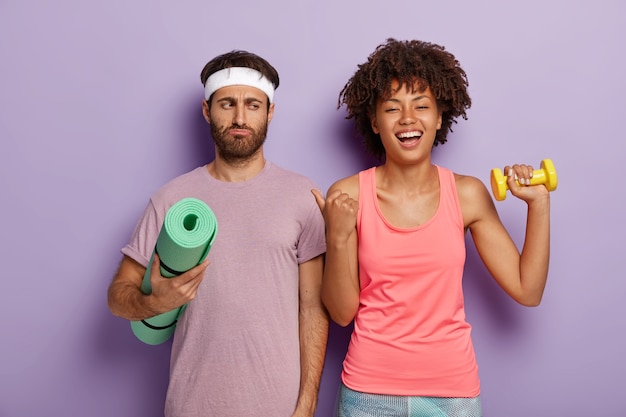 Free photo glad beautiful woman with afro haircut points thumb at husband with dissatisfied expression, have active workout together, wear sport clothes. upset guy is not eager to have fitness training