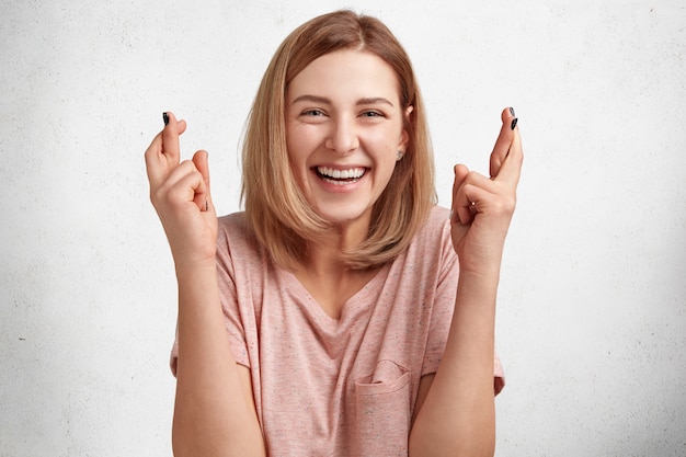 Glad beautiful young female student looks happily at camera and crosses fingers, has great desire to pass exam successfully, isolated over white concrete wall.