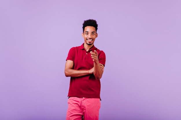 Glad black man in summer outfit posing on violet wall. carefree brunette guy expressing good emotions.