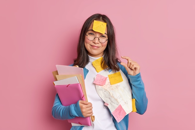 Glad brunette schoolgirl makes tiny gesture tells she needs little more time for exam preparation has sticky note stuck on forehead has deadline to finish project work.