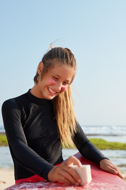 Glad Caucasian young woman in swimsuit, has toothy smile, waxes surfboard, poses against blue sky, has pleased expression