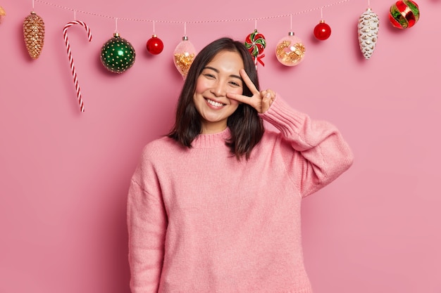 Free photo glad charming woman with toothy smile dark hair makes peace gesture expresses positive emotions dressed in casual sweater has festive mood poses