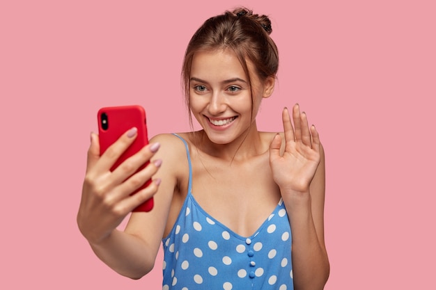 Glad European woman with pleasant smile, waves with hand  of cell phone, dressed in polka dot dress