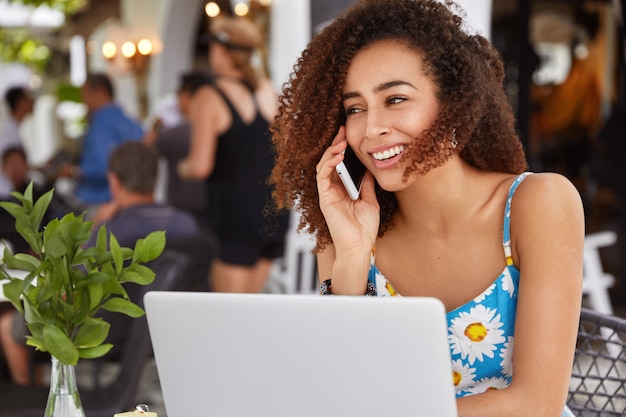 Glad female freelancer concentrated on remote job, speaks with colleague via smart phone, spends work break in cozy restaurant.