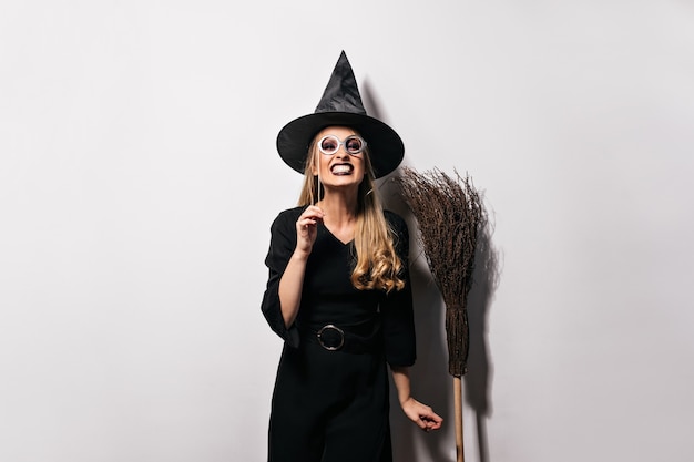 Glad female model in funny halloween costume posing . Emotional blonde girl in witch hat standing on white wall.