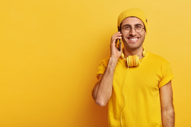 Free photo glad hipster guy with pleasant look, smiles positively, talks via smartphone, dressed in casual clothes, wears transparent glasses, discusses last fashion trends with best friend