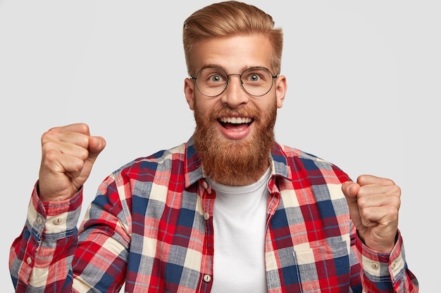 Free photo glad hipster with funny expression, clenches fists, celebrates successful day, has trendy hairstyle and ginger beard, wears bright checkered shirt, isolated over white wall. triumph concept