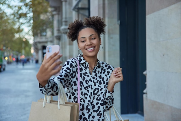 Glad millennial girl with curly hair makes selfie of herself via modern smartphone carries paper shopping bags enjoys good day dressed in fashioable clothes rejoices big sales