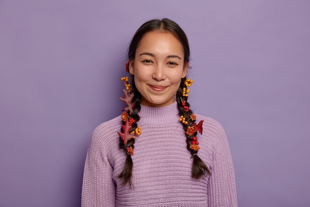 Glad natural Asian woman celebrates fall,has two plaits decorated with red autumn leaves, berries and flowers, wears purple sweater, isolated on violet wall. October time