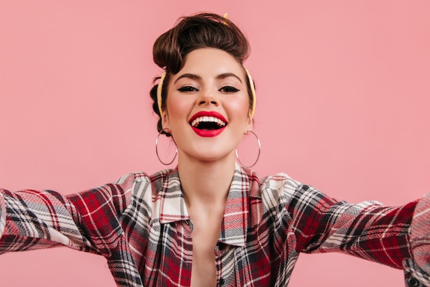 Glad pinup girl laughing on pink background. Stylish brunette woman taking selfie.