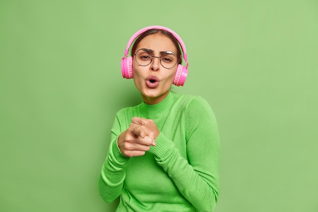 Glad playful woman sings song indicates at you enjoys favorite music in pink headphones enjoys moment wears transparent glasses and turtleneck poses against green wall