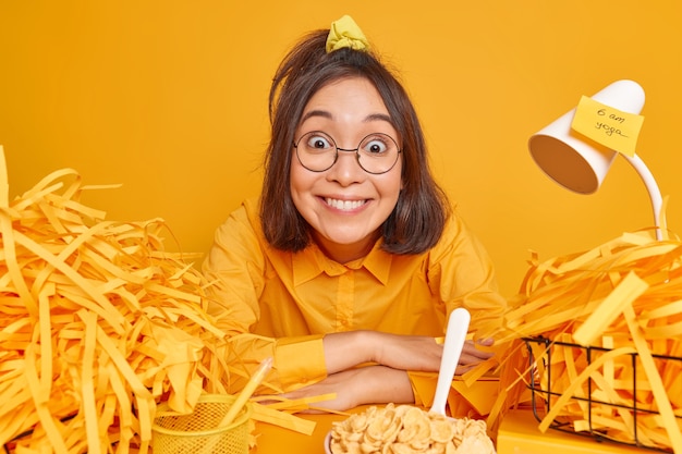 Free Photo glad positive female works at home office has busy schedule poses at workspace surrounded by cut paper after making creative project eats breakfast ready to start working sits at desktop alone