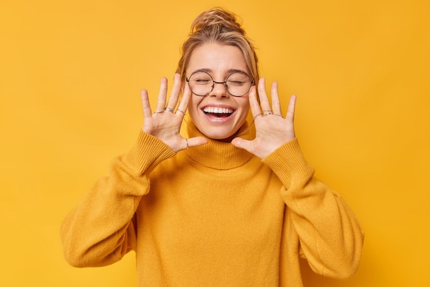 Free photo glad positive young woman keeps palms raised near face exclaims happily closes eyes wears casual jumper smiles broadly shows white teeth isolated over yellow background human emotions concept