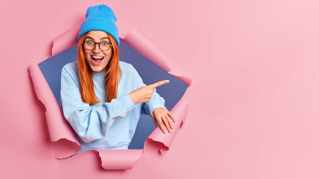 Free photo glad red haired woman with winter clothes pointing at copy space.