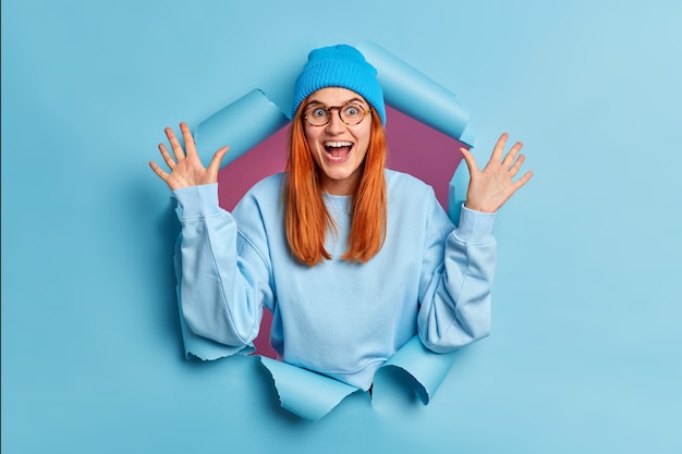 Glad redhead young woman wears optical glasses hat and jumper raises hands stands in torn paper hole excited by great news.