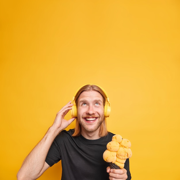 Glad relaxed ginger man looks overhead smiles broadly holds tasty ice cream listens music via headphones wears black t shirt isolated over vivid yellow wall copy space for your ptomotion