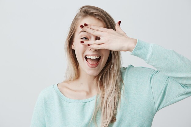 Glad smiling blonde female model hiding her face behind hand, has broad smile