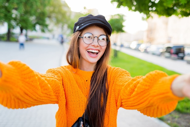 Glad smiling brunette girl in round eyeglasses and trendy orange knitted sweater making self portrait