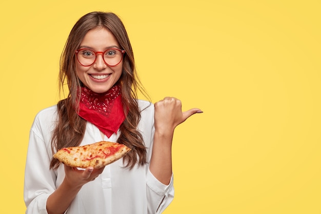 Free photo glad smiling woman holds tasty pizza, indicates with thumb aside as shows place where she bought it, advertises pizzeria, wears red bandana and white shirt, isolated over yellow  wall.