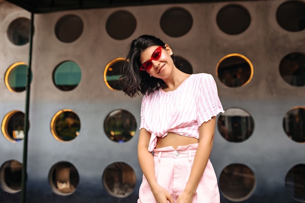 Free photo glad stylish woman looking at camera on urban background. outdoor shot of gorgeous woman in pink sunglasses posing with smile.