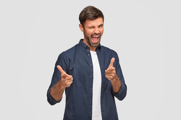 Glad unshaven male blinks with eye, opens mouth and points, feels excited, wears casual clothes, demonstrates his choice, stands against white wall. Joyful European man indoor