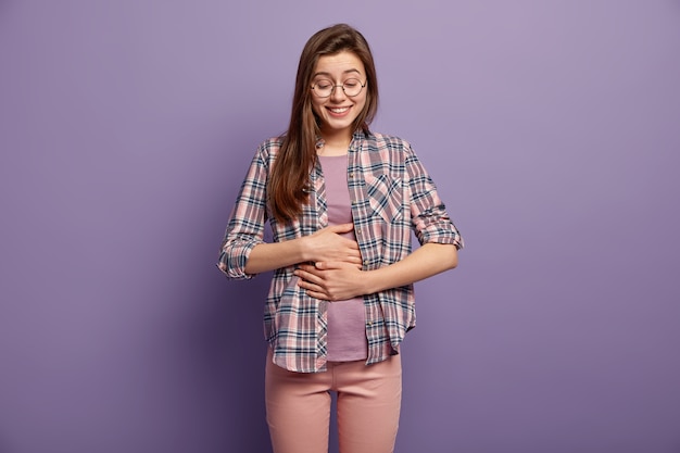 Free Photo glad woman has full stomach after delicious dinner, keeps both hands on belly, being well fed smiles positively dressed in casual clothes round spectacles isolated over purple wall. feeling of satiety