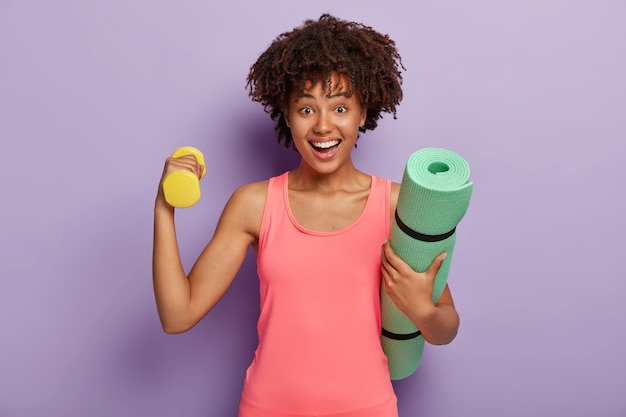 Glad woman with crisp hair, raises dumbbell for training muscles, wears pink top, carries green fitness mat