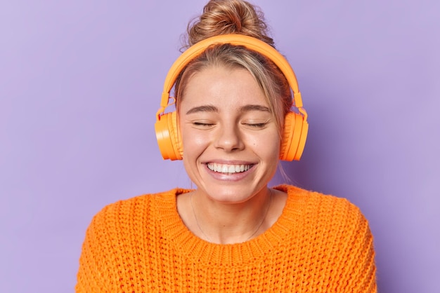 Free Photo glad young carefree woman smiles toothily keeps eyes closed dressed in warm knitted jumper listens favorite audio track via headphones poses indoor against purple background. happy emotions concept