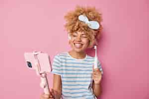 Free photo glad young woman with curly hair wears headband and casual striped t shirt records video blog holds smartphone on selfie stick gives recommendations how to care about teeth holds toothbrush